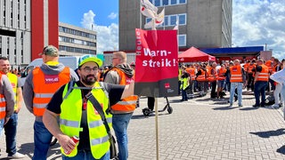 Ein Mann mit einem Schild steht vor zahlreichen Menschen in Warnwesten.