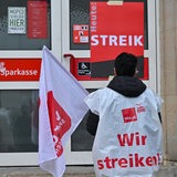 Eine Sparkassen-Mitarbeiterin steht vor der Tür einer Sparkasse. Dort steht groß: Heute: Streik.
