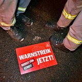 Ein Schild der IG Metall mit Aufschrift "Warnstreik Jetzt" liegt auf dem Boden. Daneben Füße in Sicherheitsschuhen.