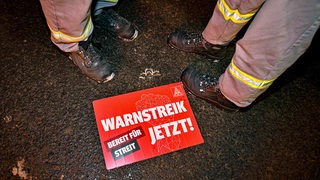 Ein Schild der IG Metall mit Aufschrift "Warnstreik Jetzt" liegt auf dem Boden. Daneben Füße in Sicherheitsschuhen.