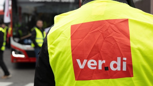 Ein Mensch in Warnweste mnit "Verdi"-Logo steht mit anderen vor einem Linienbus.