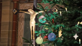 Ein Kruzifix an der Wand in einer Kirche, davor Zweige eines Weihnachtsbaums.
