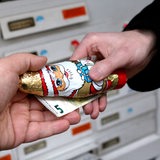 Eine Hand überreicht einer anderen Hand einen Schokoladenweihnachtsmann und einen 5-Euro-Schein vor Briefkästen an einem Hauseingang.