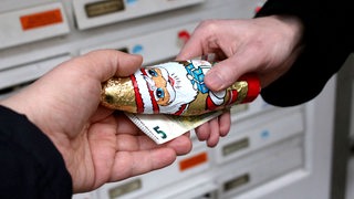 Eine Hand überreicht einer anderen Hand einen Schokoladenweihnachtsmann und einen 5-Euro-Schein vor Briefkästen an einem Hauseingang.