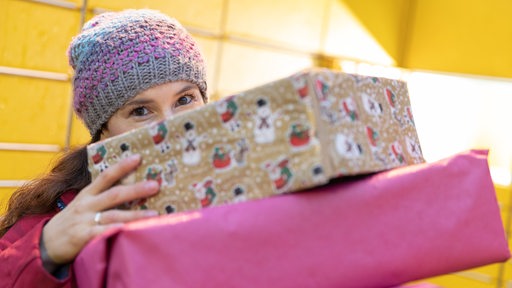Eine Frau mit Wollmütze lugt hinter einem Weihnachtspaket hervor (Symbolbild)