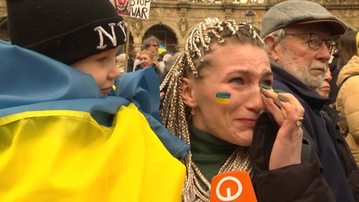 Eine weinende Frau mit Kind im Arm am Gedenktag des krieges in der Ukraine. Sie tragen eine Ukrainische Flagge.