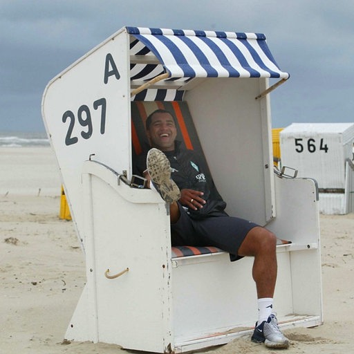 Ailton sitzt lachend in einem Werder-Trainingsanzug auf einer Strandliege. 