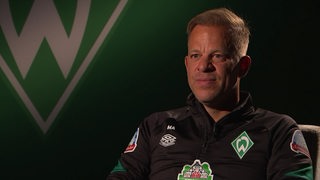 Markus Anfang sitzt in einem Sessel während des Interviews. Hinter ihm ist das Werder-Logo zu sehen.