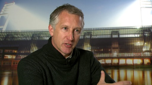 Werder-Sportchef Frank Baumann sitzt vor einem Foto des Weser-Stadions bei einem Interview.