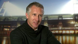 Werder-Sportchef Frank Baumann sitzt vor einem Foto des Weser-Stadions bei einem Interview.