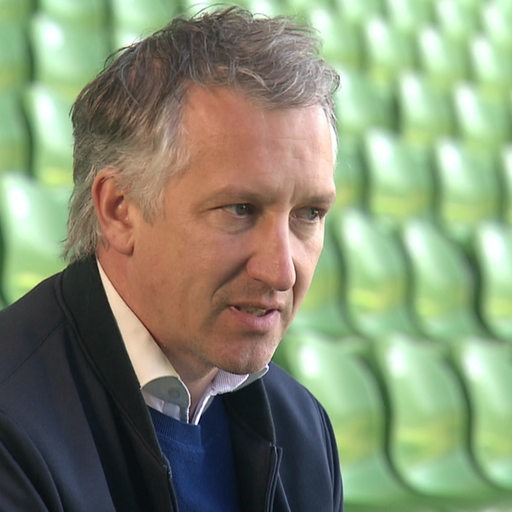 Werder-Geschäftsführer Frank Baumann sitzt auf einer leeren Tribüne auf grünen Plastiksitzen im Weser-Stadion bei einem Interview.