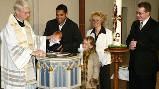 Der Probst Ansgar Lütten tauft Niklas Ailton Diegmann, während Ailton und die Eltern sowie Schwester des Taufkindes lächelnd danebenstehen.
