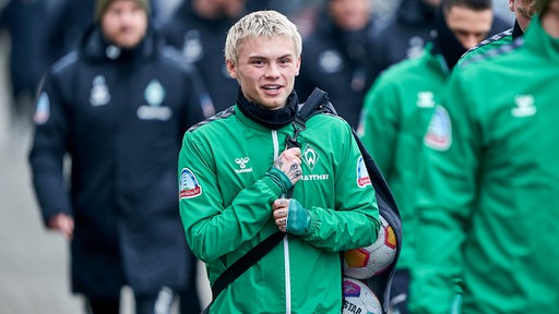 Werder-Neuzugang Isak Hansen-Aaroen läuft lächelnd und mit einem Bällesack auf dem Rücken zum Trainingsplatz.