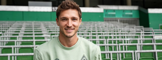 Werder-Neuzugang Niklas Stark sitzt lächelnd im Vereins-Pulli vor der Ostkurve im Weser-Stadion.