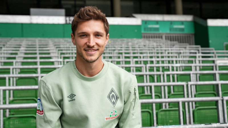 Werder-Neuzugang Niklas Stark sitzt lächelnd im Vereins-Pulli vor der Ostkurve im Weser-Stadion.