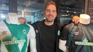 Werder-Fan-Rodrigo posiert im Fanhsop mit zwei Puppen, die Werder-Trikots tragen.