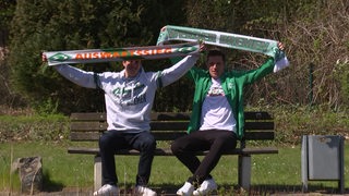 Zwei Werder-Fans aus Syke sitzen auf einer Bank und halten ihre Werder-Schals hoch.