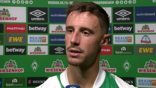 Werder-Kapitän Marco Friedl steht vor einer Werbewand beim Interview.