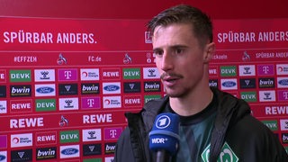 Werder-Kapitän Marco Friedl steht nach der Niederlage in Köln vor einer Werbewand beim Interview.