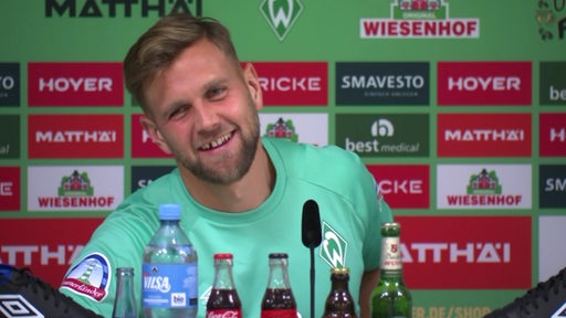 Werder-Stürmer Niclas Füllkrug legt bei einem Mediengespräch vor einer Werbewand lächelnd seinen Kopf schräg.
