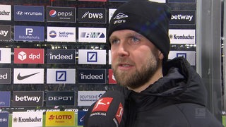 Werder-Stürmer Niclas Füllkrug mit einer schwarzen Wollmütze vor einer Werbewand beim Interview nach dem Spiel.
