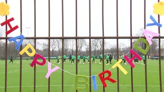 Eine bunte Girlande aus einzelnen Buchtaben formen ein "Happy Birthday" an dem Werder Trainingsplatz-Zaun.