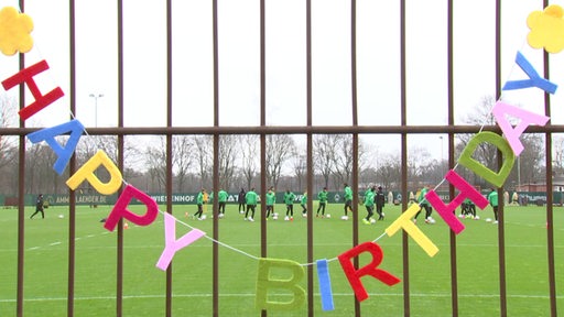 Eine bunte Girlande aus einzelnen Buchtaben formen ein "Happy Birthday" an dem Werder Trainingsplatz-Zaun.