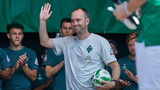 Werder-Trainer Ole Werner winkt lächelnd mit einem Ball unter dem Arm auf der Bühne beim "Tag der Fans".