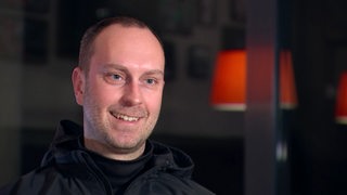 Werder-Trainer Ole Werner sitzt in seiner schwarzen Trainingsjacke lächelnd bei einem Interview.