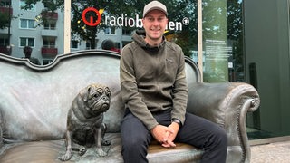Werder-Trainer Ole Werner sitzt vor seinem Besuch bei Radio Bremen auf dem Bronze-Sofa von Loriot neben dem Mops.