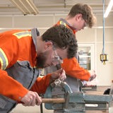 Zwei Azubis in orangfarbenen Overalls arbeiten in einer Werkstatt.