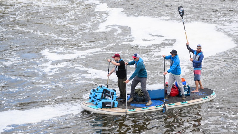 Vier Männer stehen im Fluss auf einem SUP und paddeln.