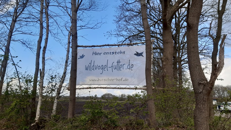Ein Schild mit der Aufschrift "Hier entsteht wildvogel-futter.de" hängt zwischen Bäumen.