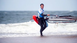 Windsurfer Lennart Neubauer trägt mit breitem Strahlen sein Bord ans Strandufer von Sylt nach dem erfolgreichen Freestyle-Wettbewerb auf Sylt.