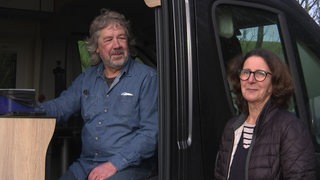 In einem Camper-Van sitzt ein Mann, der zu seiner Frau blickt, die neben dem Auto steht.