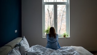 Eine Frau sitzt auf dem Bett und schaut aus dem Fenster (Symbolbild)