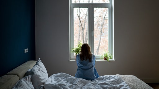 Eine Frau sitzt auf dem Bett und schaut aus dem Fenster (Symbolbild)