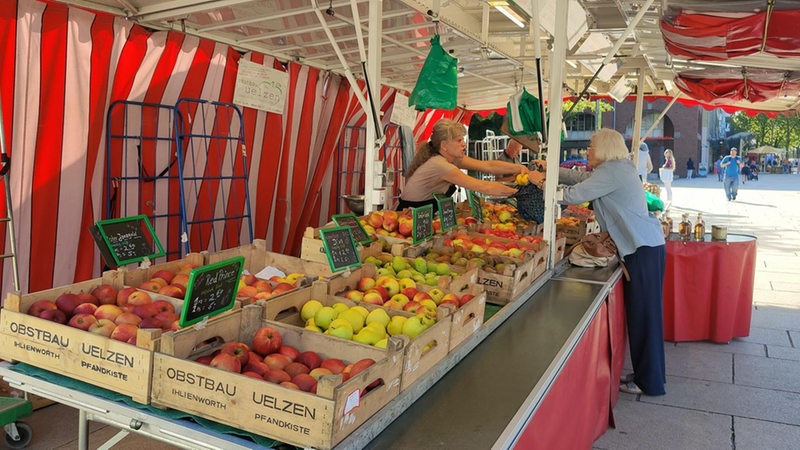 Auf einem Wochenmarkt kauft eine Kundin an einem Obststand Äpfel ein.