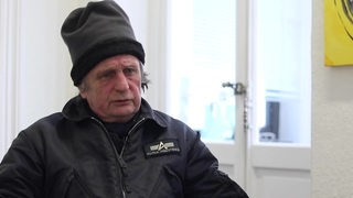 Ein Mann sitzt in einem weißen Raum, hinter ihm eine Tür mit Fenstern, er trägt eine hohe Mütze und eine Jacke