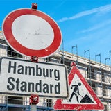 Baustellenschild mit Zusatzschild mit der Aufschrift: "Hamburg Standard" auf Baustelle vor einem Mehrfamilenhaus-Rohbau mit Baugerüsten (Symbolbild)