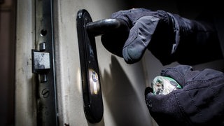 Eine Hand im Handschuh greift nach einem Türgriff einer schweren Tür, die andere Hand hält eine Taschenlampe.