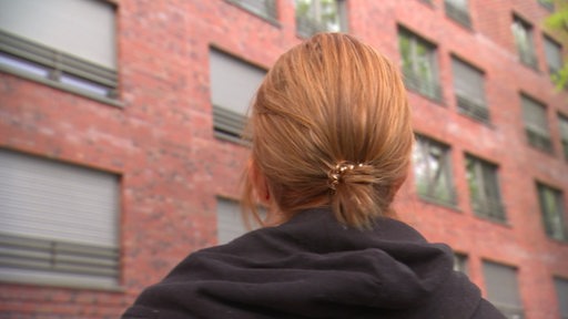 Der Hinterkopf einer Frau mit roten Haaren ist zu sehen, die Frau steht vor einem Gebäude.