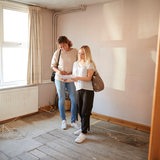Zwei junge Frauen stehen in einer leeren Wohnung und schauen auf einen Plan.