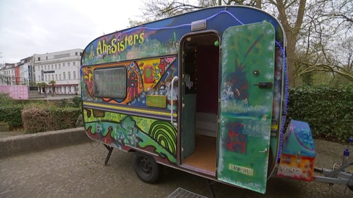 Ein mit Graffiti besprühter Wohnwagen, der zu einem begehbaren Kunstprojekt umgewandelt wurde, steht in einem Innenhof.