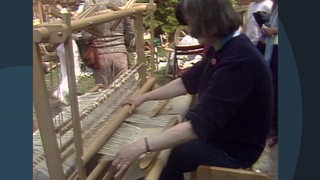 Eine Frau sitzt an einem Webstuhl und webt Wolle zu einem Tuch. 