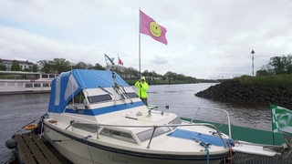 Auf einem Boot ist eine Person zu sehen, die eine Flagge mit einem Smiley drauf in den Händen hält. 