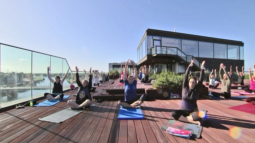 Auf einer Dachterrasse machen mehrere Personen bei einem Yoga-Kurs mit.