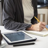 Eine Frau zeichnet mit einem Bleistift auf einen Zeichenblock - als Vorlage dient ein Bild auf einem Tablet.
