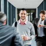 In einem Büroflur sieht man einen ruhigen Angestellten mit Kaffeebecher neben unscharfen Kollegen in Hektik.