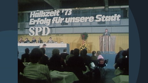 Menschen sitzen hinter einem Tisch auf einem Podium, davor sitzen Zuschauer. An der Wand steht "Halbzeit '73. Erfolg für unsere Stadt. SPD"